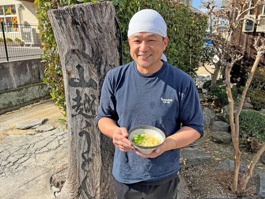 看板メニューの「釜玉うどん」を手に笑顔を見せる３代目社長の山越伸一さん