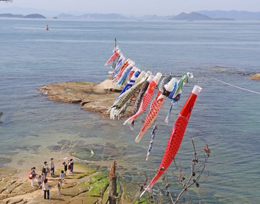 潮風を受け、青い海や空を背景に泳ぐこいのぼりを眺める家族連れ＝香川県小豆郡土庄町、戸形公民館前