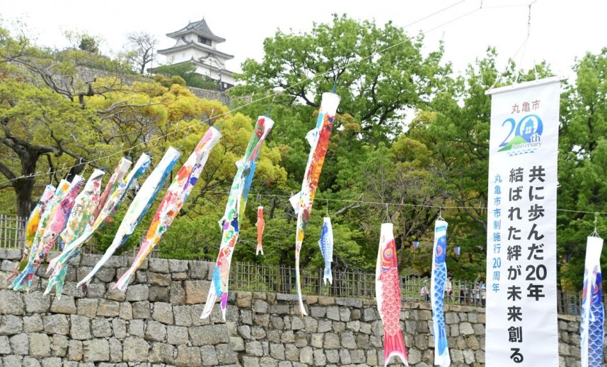 丸亀城の堀の上を悠々と泳ぐこいのぼり。市制施行２０周年記念の垂れ幕も一緒につるされている＝香川県丸亀市一番丁