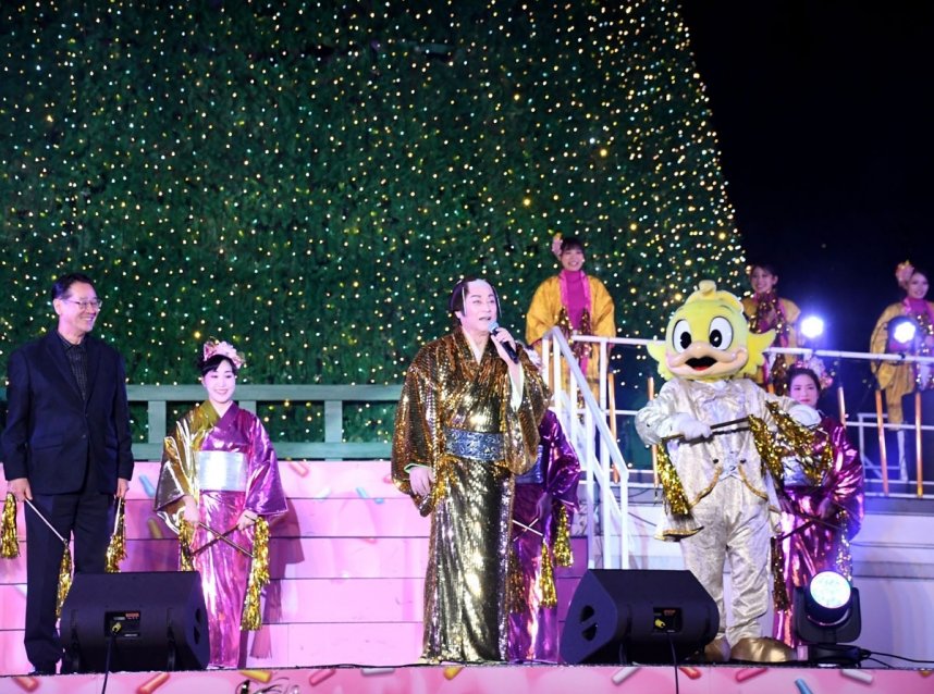 金色に点灯したクリスマスツリーの前で観客に感謝を伝える松平さん（中央）＝香川県丸亀市綾歌町、ニューレオマワールド